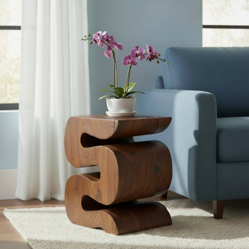 Haussmann Big Wave solid wood end table in mocha finish, 12 x 14 x 18 inches, sculptural S-curve design with natural knots and fine surface checks in the grain, styled in a living room beside a medium blue sofa with a white ceramic bowl and purple orchid, near a window with white sheer curtains and natural sunlight.